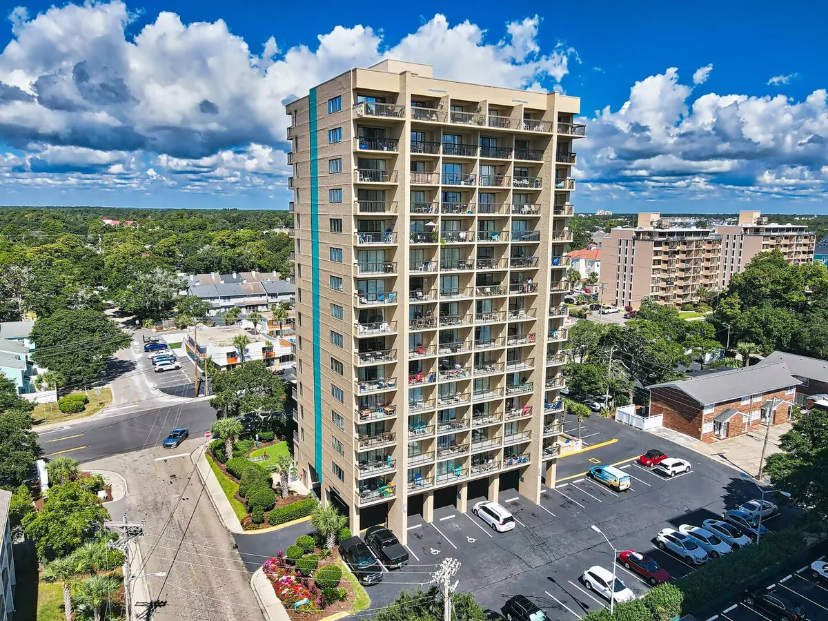 A modern high-rise building with balconies adorned with plants, surrounded by lush greenery and a welcoming parking area. The setting radiates a sense of peaceful residential living in a vibrant neighborhood.