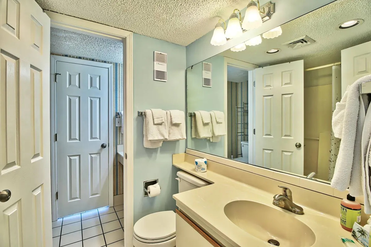 A bright bathroom featuring a mirror, modern sink, soft towels, and elegant lighting fixtures with doors in the background. The clean design suggests comfort and privacy for daily routines.