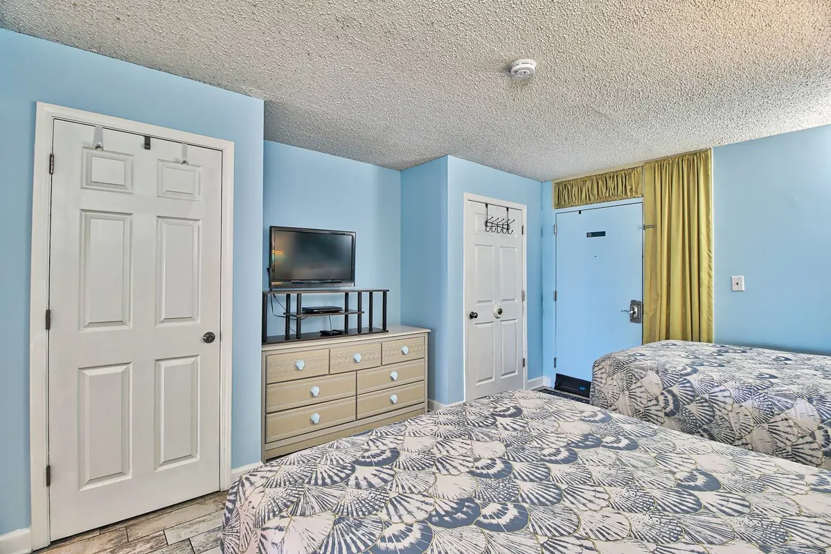 A casual, relaxed bedroom environment with two patterned beds, a television on a stand, dresser for storage, and curtain-covered window. The layout suggests comfort, space for belongings, and entertainment options.