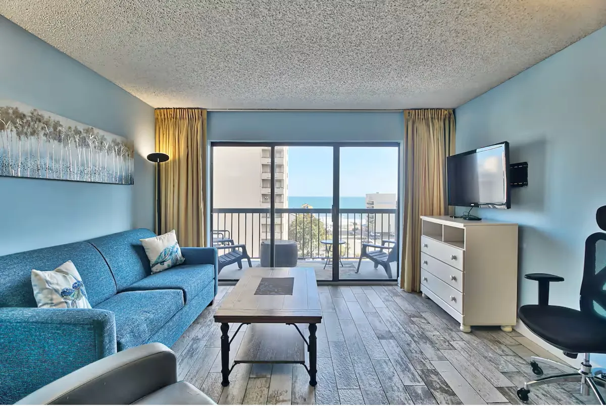 A spacious living area featuring a plush sofa and coffee table, with large glass doors opening directly onto a private balcony. The room is bright and inviting, with sunlight streaming in and the ocean visible in the background. The setup encourages both relaxation and socializing, with the television and seating oriented for group gatherings or quiet evenings. The seamless connection between the living space and the balcony gives a sense of indoor-outdoor living.