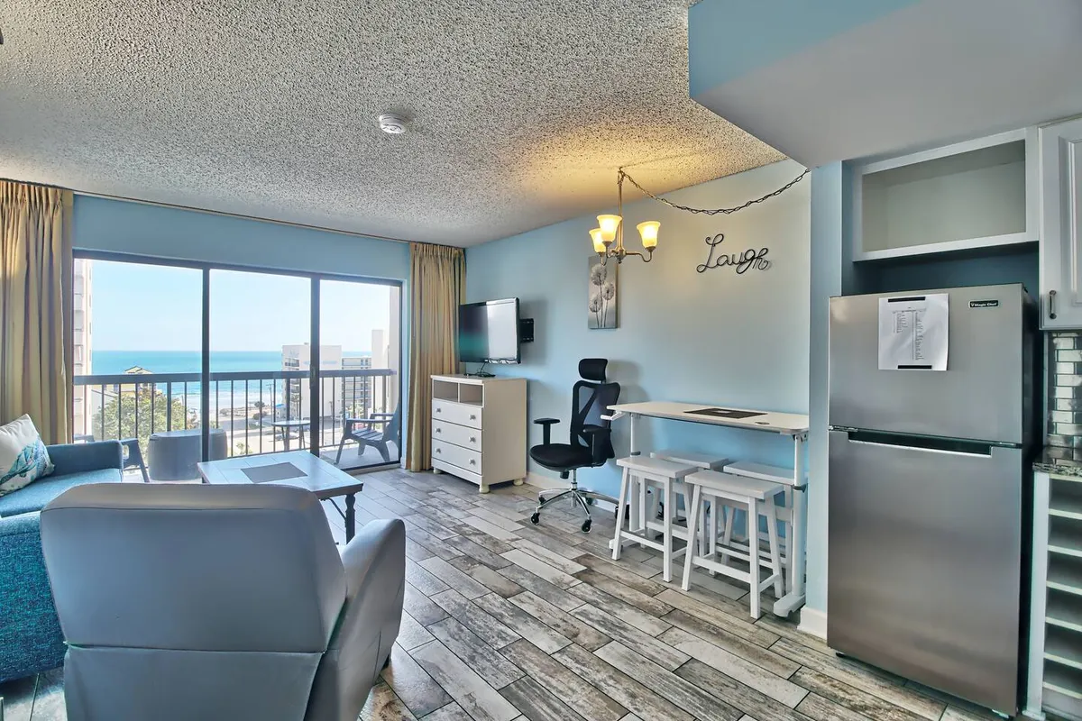 A bright, spacious living area with a large window overlooking the ocean. The room features a blue sofa, a dining table with chairs, and a refrigerator, creating a seamless blend of comfort and functionality. Natural light fills the space, emphasizing the welcoming, home-like atmosphere.