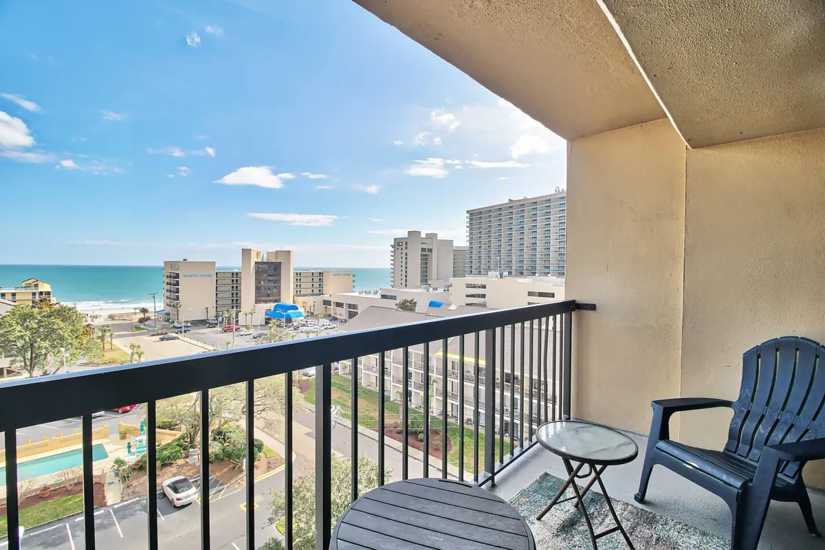 A private balcony set for two, with comfortable outdoor chairs and a small table, directly overlooking the shimmering ocean. The balcony setting is serene and inviting, perfect for morning coffee, sunset toasts, or intimate moments. Distant hotels and beach scenery anchor the scene in Myrtle Beach’s iconic landscape.