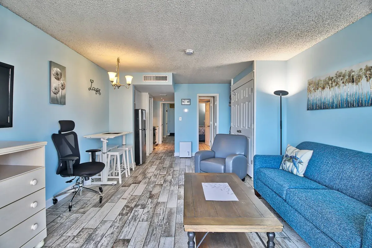 An inviting living area centered on a blue couch, wooden coffee table, and modern chair, with wall art and a glimpse of the kitchen. The open concept provides ample space for relaxing, socializing, or even movie nights.