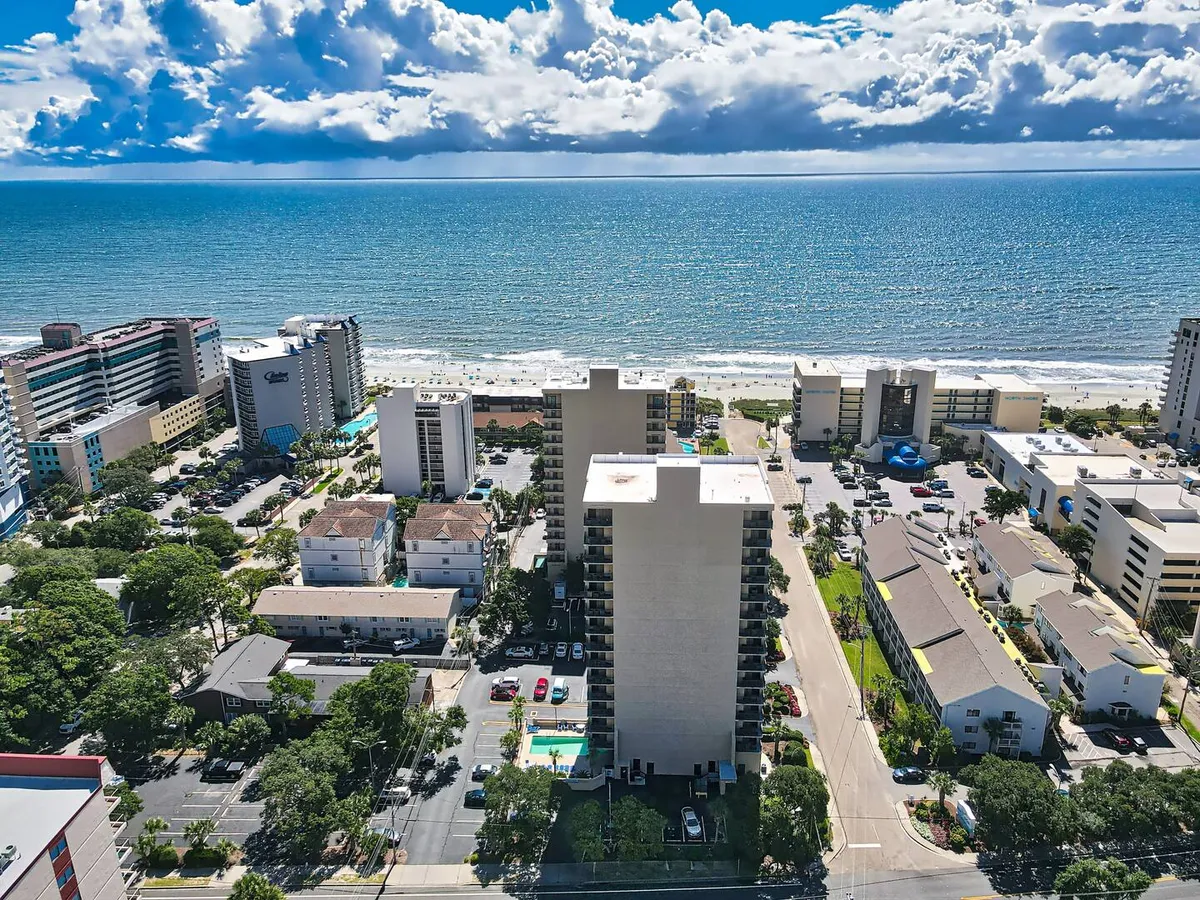 A sweeping ocean view with high-rise buildings, lush landscaping, and a parking area in the foreground. The scene suggests proximity to both the beach and local attractions, capturing the vibrancy of the surrounding area and easy access to recreation.