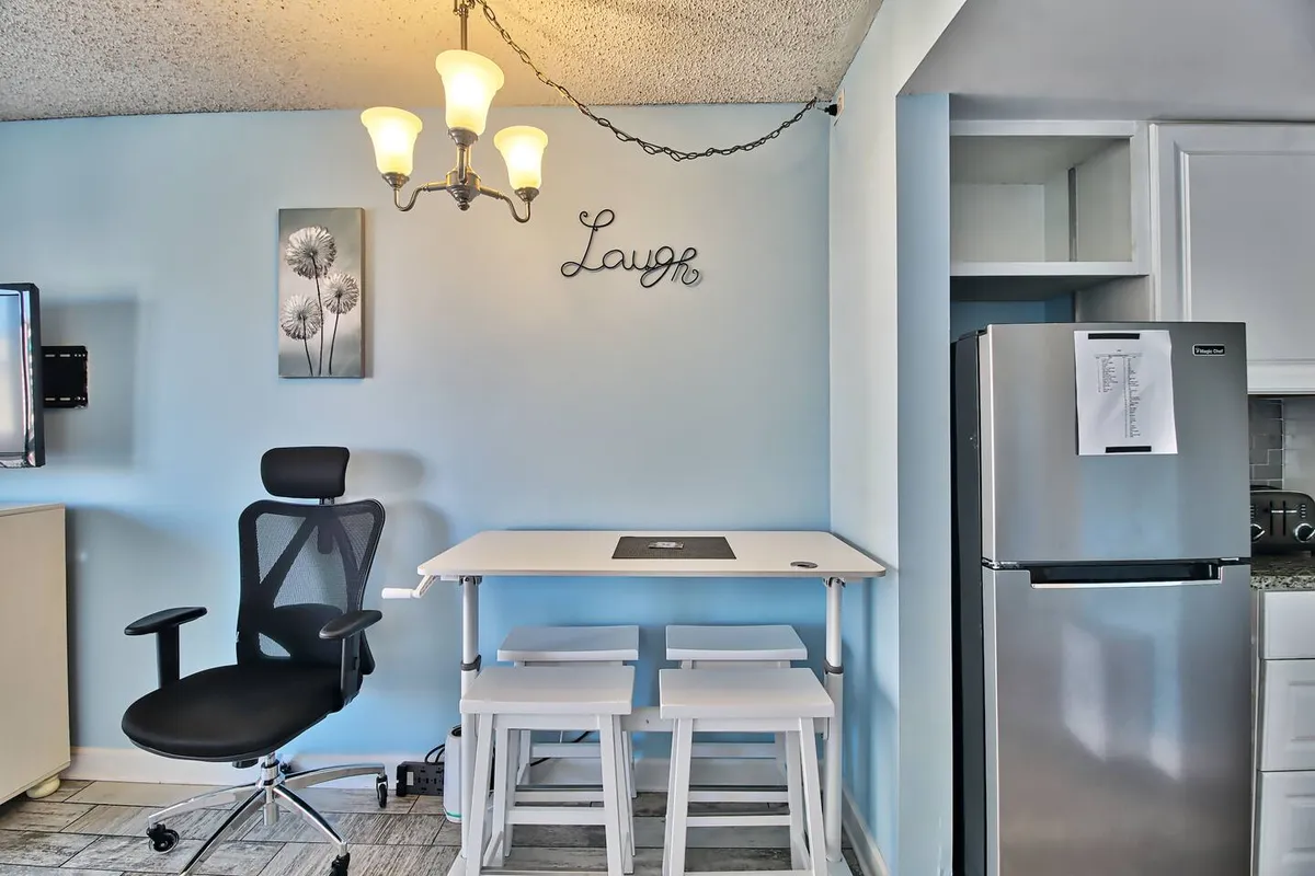 A cheerful dining nook with a chandelier overhead, a table surrounded by comfortable chairs, and playful 'Laugh' wall decor. The refrigerator is visible in the background, connecting this area seamlessly with the kitchen and reinforcing a sense of togetherness and casual dining.