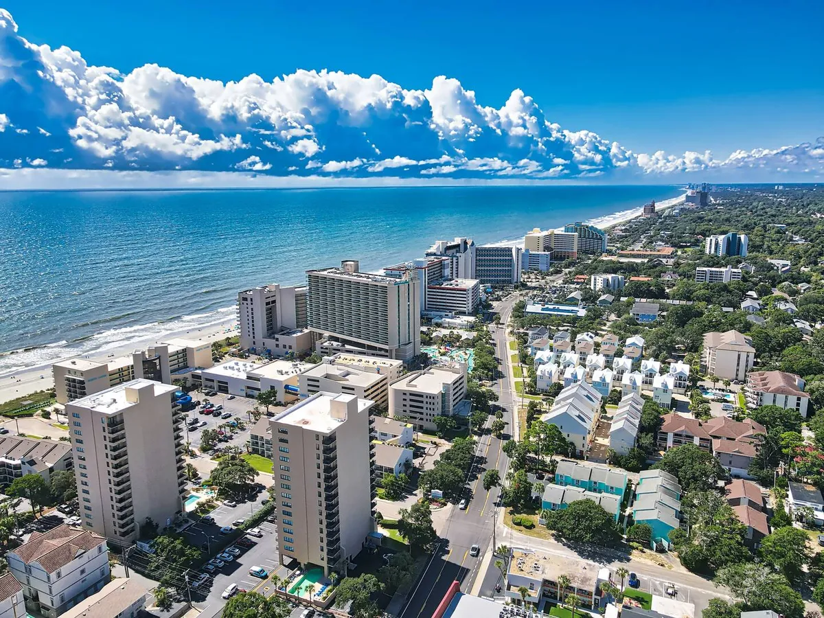 A sweeping view of the Atlantic Ocean, with buildings, the sandy beach, and a road visible in the foreground. The sky is adorned with fluffy clouds, and the overall scene is vibrant and energetic, showcasing the lively oceanfront environment.