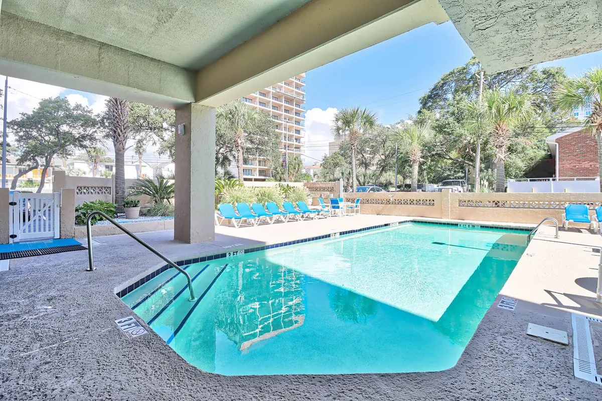 The image features a sparkling swimming pool surrounded by blue lounge chairs and palm trees, evoking a laid-back, social atmosphere. The pool area is bathed in sunlight, suggesting a perfect spot for friends to relax, sunbathe, or gather before heading out for the night.